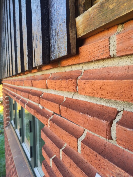 Röd tegelvägg med synliga sprickor och delade fogar ovanför en husfasad med träpanel och fönster.