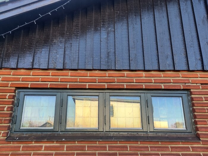 Fönster med tegelvägg och mörk träpanel ovanför, reflekterande glas. Alla skift har släppt och det översta går in under panelen.