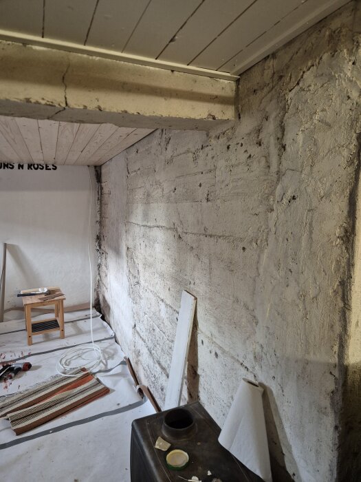 Ojämn innervägg i källare med gammal puts, träpanel i taket, verktyg och material utspridda på golvet bredvid väggen.