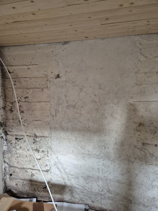Ojämn källarvägg med delvis putsad yta och synliga träpaneler i taket. Kabel hänger längs väggen.