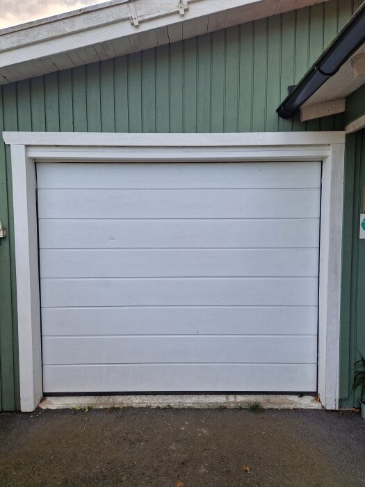 Vit elektrisk takskjutport i sektioner på ett grönt garage med behov av gångdörr för bättre isolering och passage vid strömavbrott.