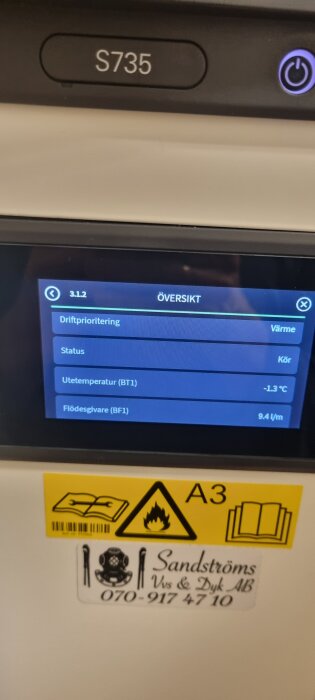 Display på en Nibe S735 värmepump som visar driftinformation, inklusive temperatur och flödesvärden, samt varningar och en företagsdekal.