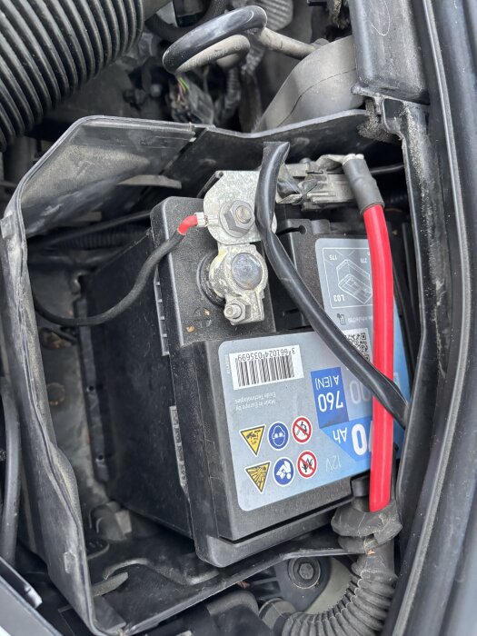 Bilbatteri med minuspol och pluspol markerade, anslutningar för laddning. Fråga om jordning på plus- eller minuspolen.