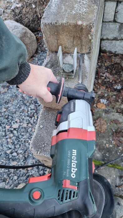 Person använder kraftfull borrhammare från Metabo för att arbeta på en betongmur, omgivna av stenar och grus.