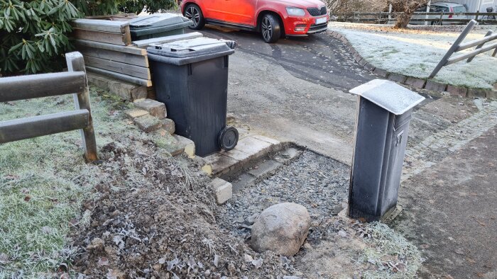 Grusig yta bredvid en liten mur och soptunna vid en uppfart. En röd bil är parkerad i bakgrunden på en frostig morgon.