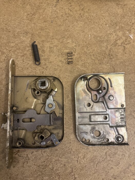 Isärplockat dörrlås med synliga fjädrar och mekanismer på en träyta, visar delar av låskistan och innanmätet i metall.