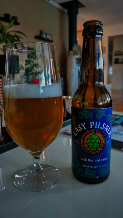 Ett glas med öl bredvid en flaska "Easy Pilsner" står på ett bord i ett hem.