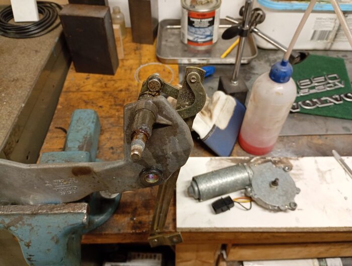 Motor och fäste för en bakrutetorkare på en arbetsbänk, omgiven av verktyg och smörjmedel, mot en rörig bakgrund i en verkstadsmiljö.