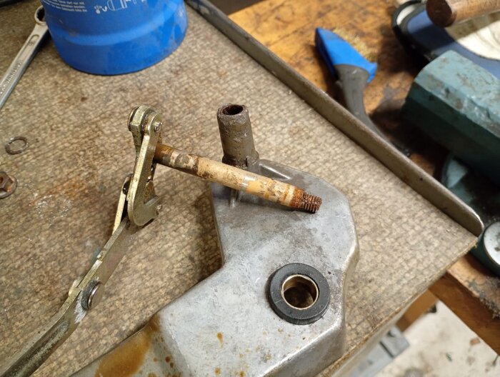 Rostig axel och fäste för bakrutetorkarmotor, delvis demonterad, på en arbetsbänk med verktyg i bakgrunden.