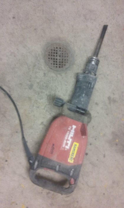 Röd och svart bilmaskin med borr bredvid golvbrunn på betonggolv i garage, använd vid byte av golvbrunnar.