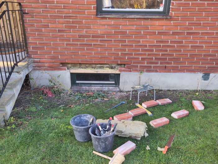 Murverk med fönster, armering över fönsteröppning och byggredskap på gräsmattan framför en tegelvägg.