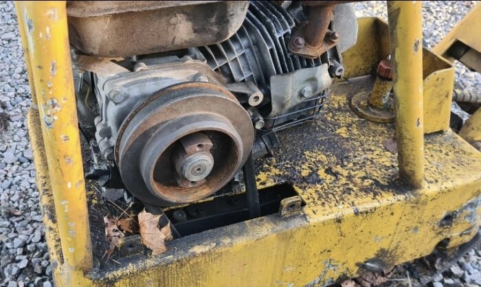 Gul, rostig maskindel på mark av grus. Möjlig motor eller kopplingsdel utan synbara märkningar eller typ-skylt. Lämplig för identifiering.