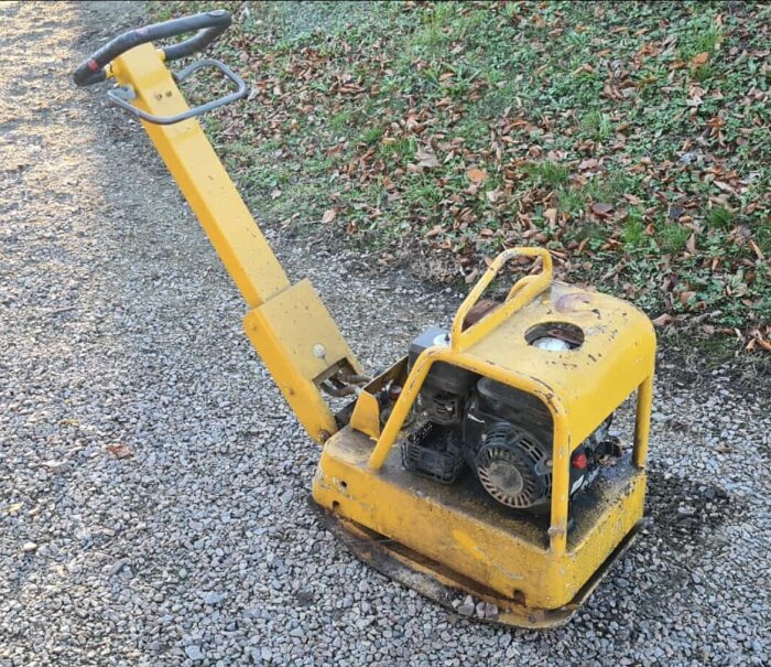 Gul markvibrator utan synlig märkning eller typ-skylt, stående på grusväg nära grönskande bakgrund.