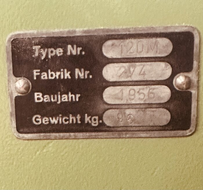 Metallskylt på grön yta med text som anger typnummer, fabriksnummer, byggår och vikt för en maskin. Skyltens hörn är fästade med skruvar.