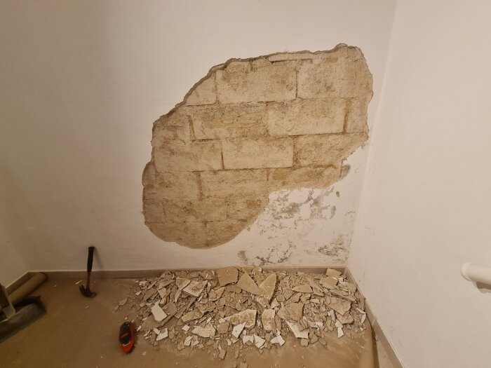 Källarvägg med bortslagen puts, blottlagda tegelstenar och krossat material på golvet. Hammare synlig på golvet bredvid.