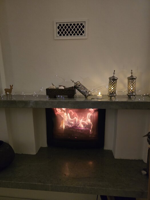 Eld brinner i en kamin under en ventil. Små dekorationer och lampor pryder hyllan ovanför kaminen.