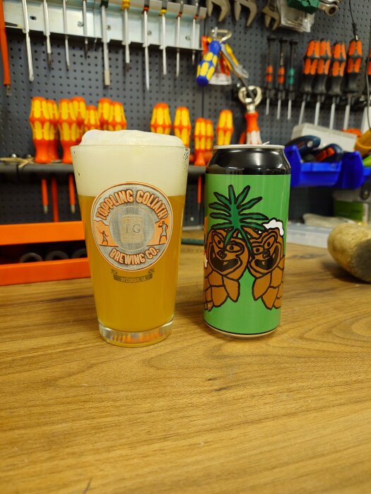 Ölglas och ölburk med Omnipollo-motiv på arbetsbänk, omgiven av verktyg och orange skruvmejslar i bakgrunden.