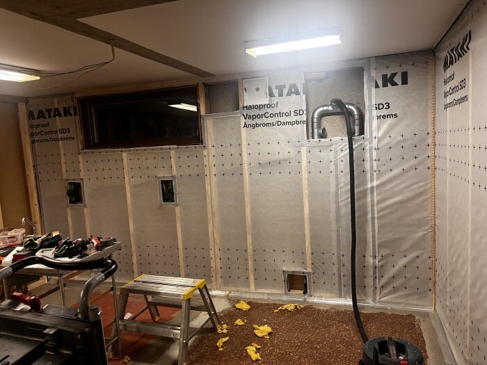 Källarväggar med undertrycksventilation och plastisolering, byggverktyg på en arbetsbänk, stege och dammsugare på golvet.