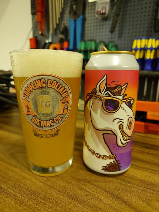 Ölglas med "Toppling Goliath Brewing Co." och en färgglad ölburk står på en träbänk i garage. Verktyg syns i bakgrunden.