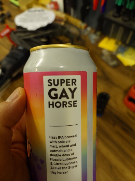 Burk med öl av märket "Super Gay Horse", en hazy IPA, hålls i handen. I bakgrunden syns verktyg i ett garage.
