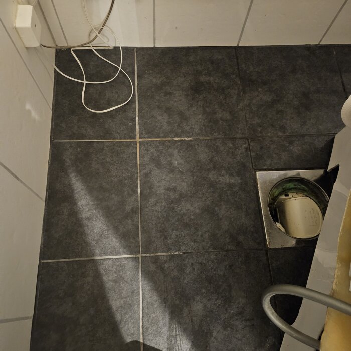 Golvbrunn i badrum med mörkgrå klinker, rensat och reparerat rör synligt, vit kabel längs väggen, del av vägg syns.