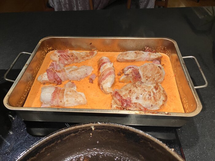 Baconlindad kyckling i en ugnsform, täckt med en röd sås gjord av ajvar och grädde.