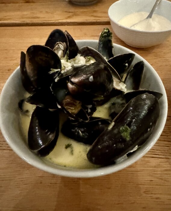 Skål med blåmusslor i sås serverad på träbord, med en vit skål innehållande sås och en sked i bakgrunden.