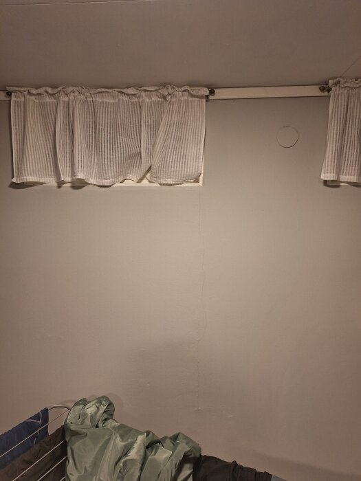 Spricka i en vitmålad källarvägg från fönster till golv, med vit gardin ovanför och torkställning nedanför.