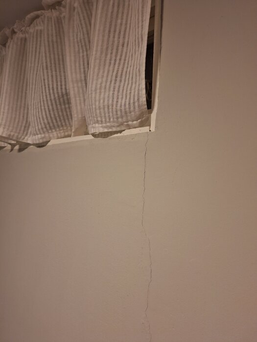 Spricka i källarvägg från hörnet av ett fönster ner till golvet, bredvid ett vitt randigt draperi.