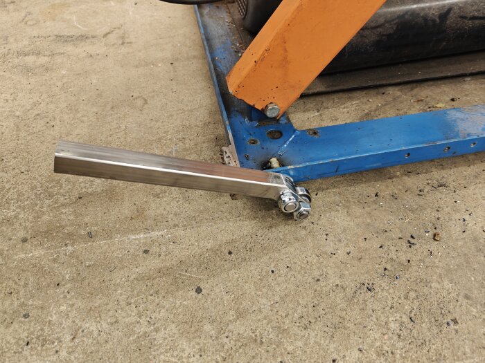 Ett svetsat metallstöd med kullager och fyrkantsrör är fastsatt på en färgad metallram liggande på golvet.