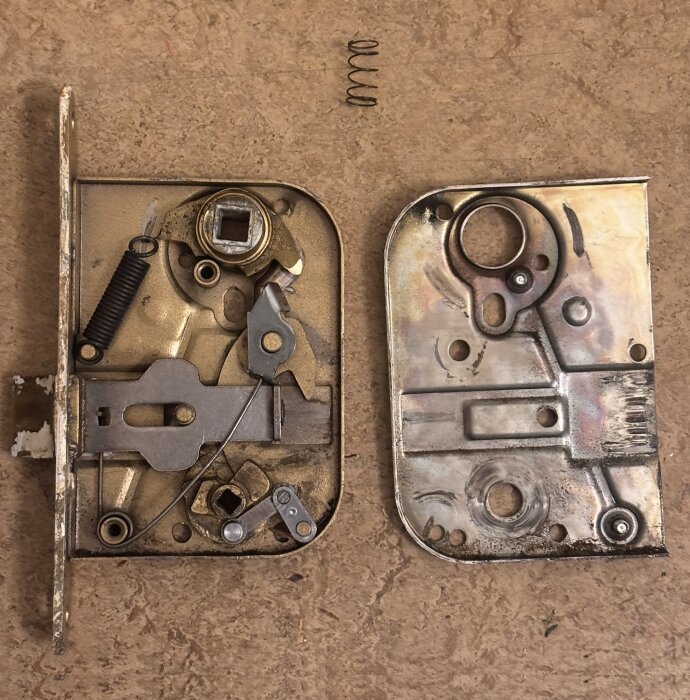 Isärplockat dörrlås med interna komponenter och en lös fjäder ovanpå, visar delarnas placering och konstruktion.