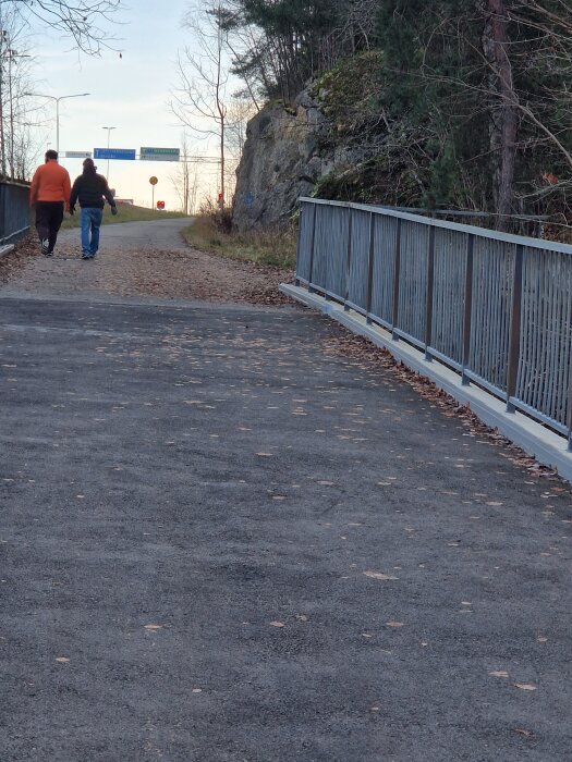 Två personer promenerar på en asfalterad gångväg bredvid ett staket, omgivna av träd och klippor under hösten.