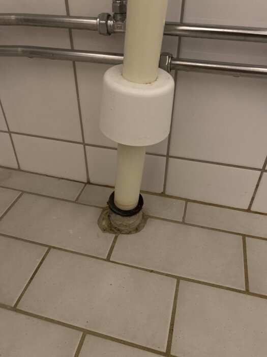 Rör som sticker upp från klinkergolv med tätningsmanchetter, nära vit kakelvägg och kromade rör.
