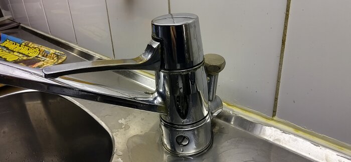 Äldre köksblandare, troligen daterad till 90-talet, med okänd tillverkare installerad i diskbänk och omgiven av en metallisk yta och kakelvägg.