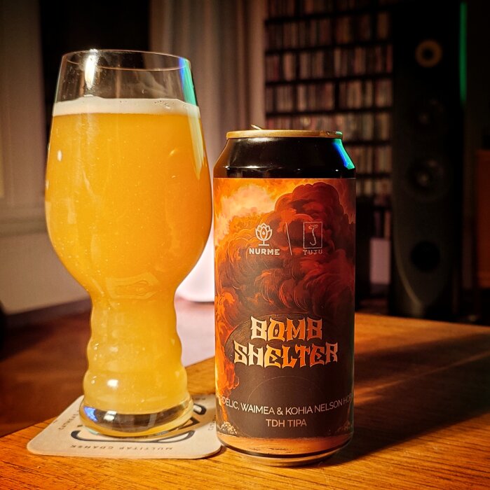 Ett glas med ljus, grumlig dryck bredvid en ölburk med etiketten "Bomb Shelter" på ett träbord i ett rum.