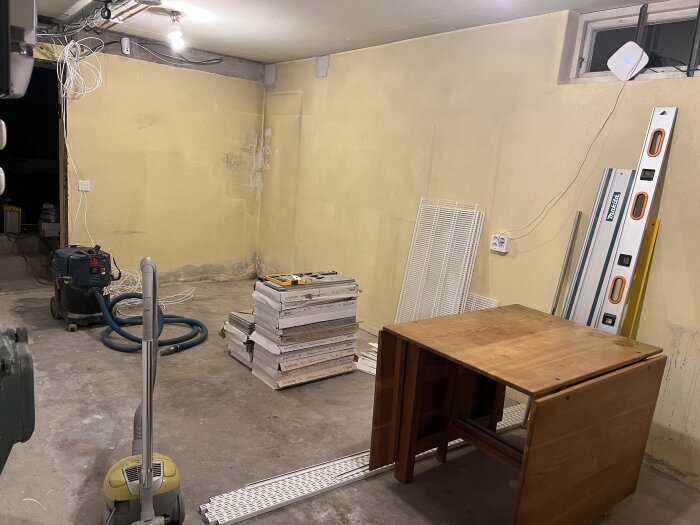 Rum med gul putsad vägg, staplade plankor, arbetsbord, dammsugare och byggverktyg. Källarfönster i väggen, vita kabeldragningar syns.