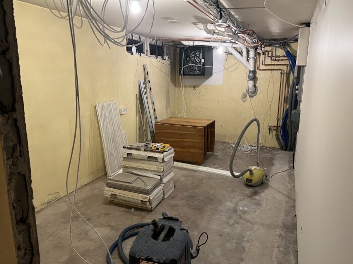 Rum under renovering med utspridda kablar, verktyg, ett ekbord och dammsugare på ett betonggolv. Kartonger och hyllor står staplade längs väggarna.