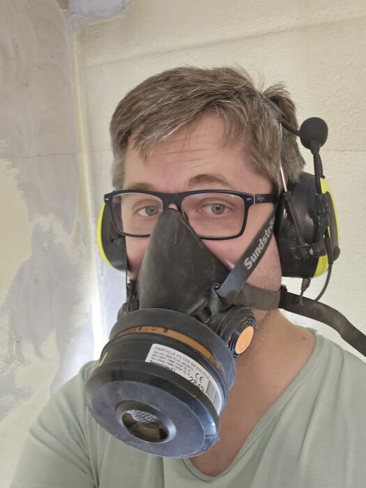 Person med skyddsglasögon och andningsmask arbetar i källare, förbereder för betongslipning. Bakgrund visar en vägg i bearbetning.