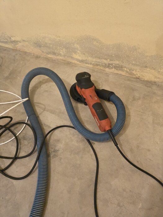 Betongslip Hilti DGH 130 med dammhuva ligger på golvet intill en vägg, omgiven av blått dammsugarfilter och kablar.