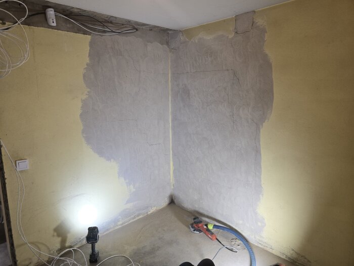 Vägg i renoveringsprojekt, delvis slipad från gul färg till bar puts; betongslip och belysning synliga på golvet.