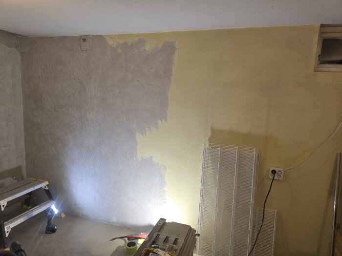 Vägg i källare under slipning, med ljusficklampa, stege och flera verktyg. Ena sidan nyputsad, andra gulmålad. Elektriskt uttag syns.