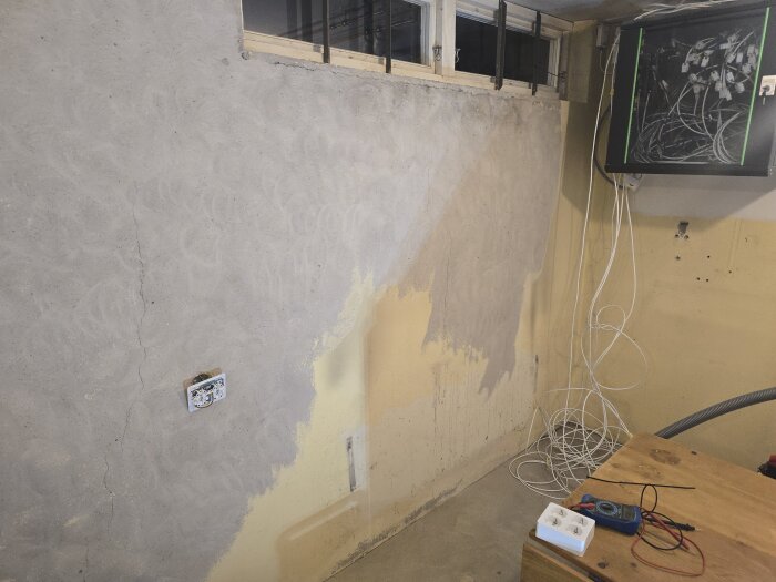 Vägg i ett gym/hobbyrum håller på att slipas, med spår av borttagen färg. Fönster upptill och elutrustning samt kablar till höger.