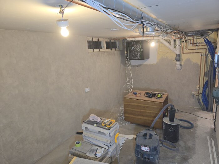 Källarrum under renovering, med betongväggar, synliga rör och kablar, dammsugare med cyklonfilter och verktyg placerade på en låda och staplade kakel.
