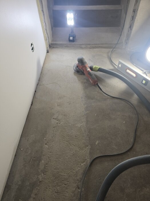 Betonggolv slipas med en dammsugaransluten vinkelslipmaskin; ljus lyser på den delvis slipade ytan i ett källarrum.