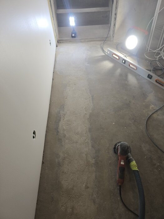 Betonggolv i ett renoveringsprojekt med en slipmaskin i förgrunden, lysande arbetslampor och en vägg med hål för elinstallationer.
