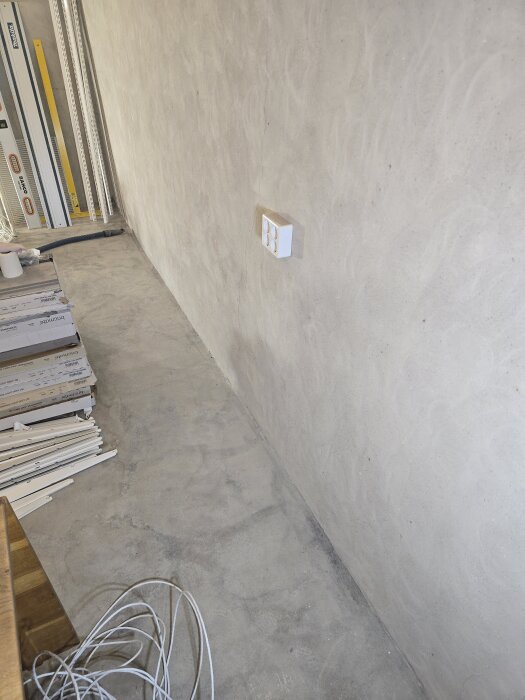 Betongvägg med ett nytt eluttag i ett rum under renovering. Golvet är slipat och det ligger verktyg och kablar på marken.