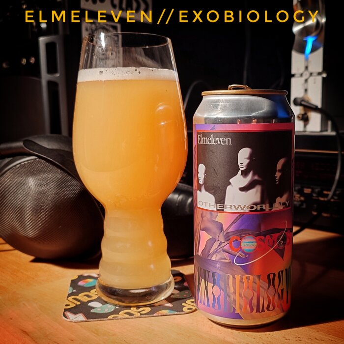 Ölglas med grumlig dryck bredvid en burk med etiketten "Elmeleven Otherworldly" och futuristisk konst står på ett svart underlägg.
