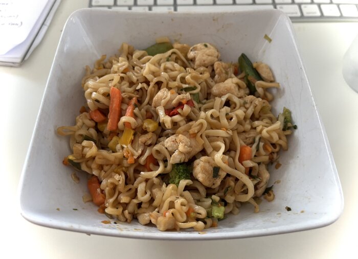 Skål med snabbnudlar, stekt kyckling och wokgrönsaker, serverad på ett bord med laptop-tangentbord i bakgrunden.