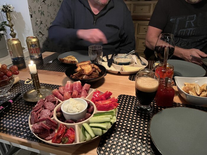 Assorterade tapas på bord med kött, grönsaker, dippsåser, bröd och dryck. Två personer sitter runt bordet.
