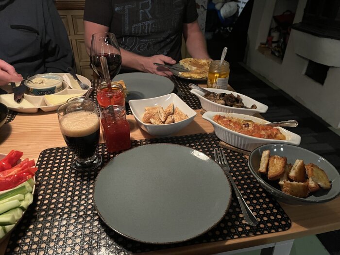Tapasmåltid med olika rätter, inklusive hummus, bröd, grönsaker, såser och ett glas dryck på ett matbord.